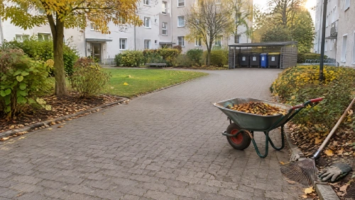Gepflasterter Innenhof einer Wohnanlage mit Schubkarre voller Laub, Rechen und Müllcontainern